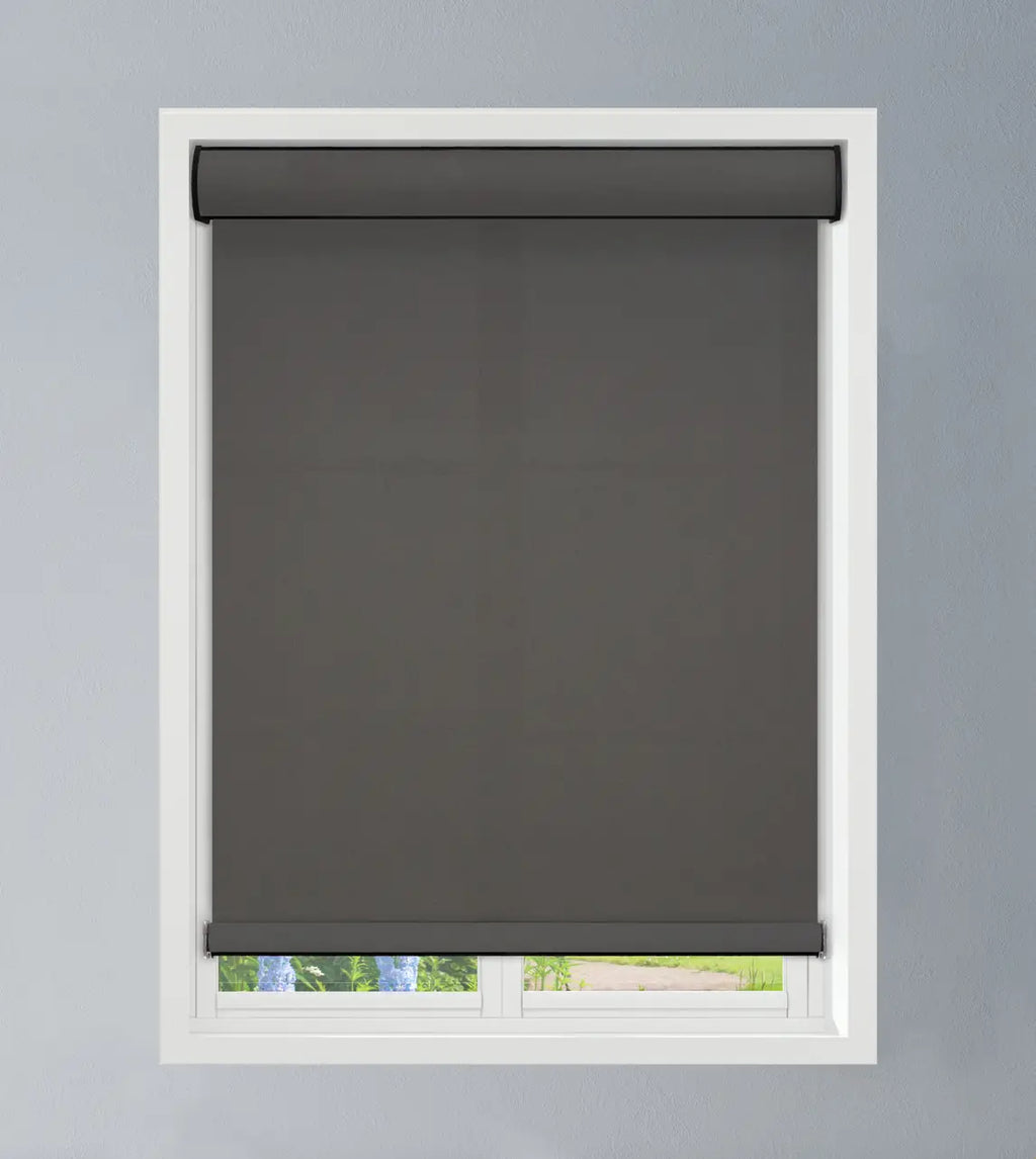 Dark gray roller shade mounted above a white-framed window overlooking a green outdoor view, shade in raised position